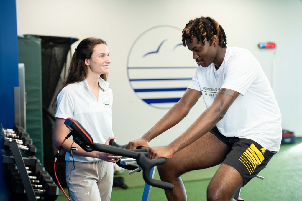 a therapist oversees a patient riding a stationary bike in a clinic setting.