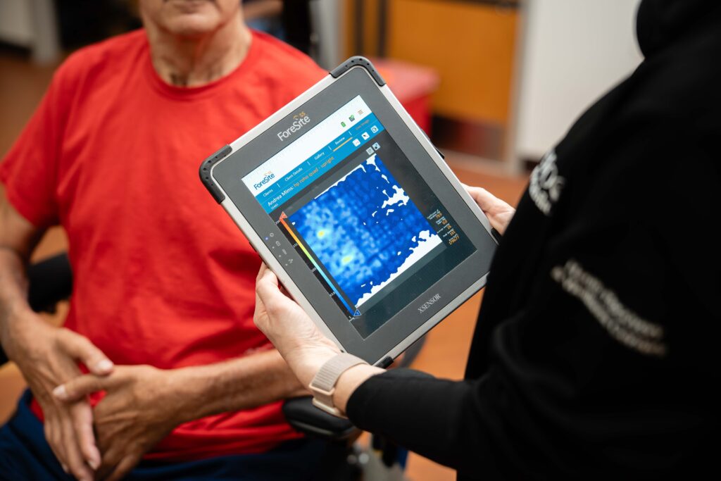 physical therapists holds an ipad while conducting pressure mapping for a patient sitting in a wheelchair at Brooks' Seating and Mobility Clinic.