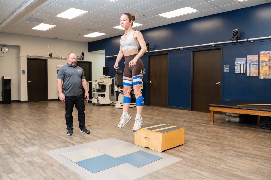 A female athlete jumps in the air while a sports therapist observes her movements.
