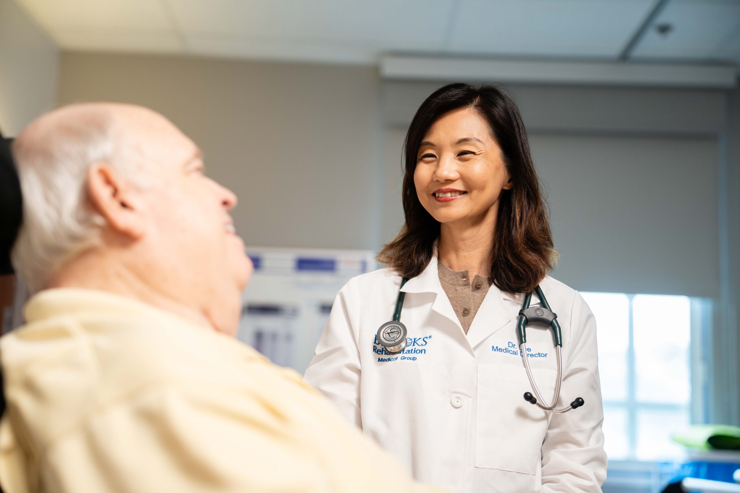 smiling woman PM&R doctor talks with an elderly male patient in their hospital room.