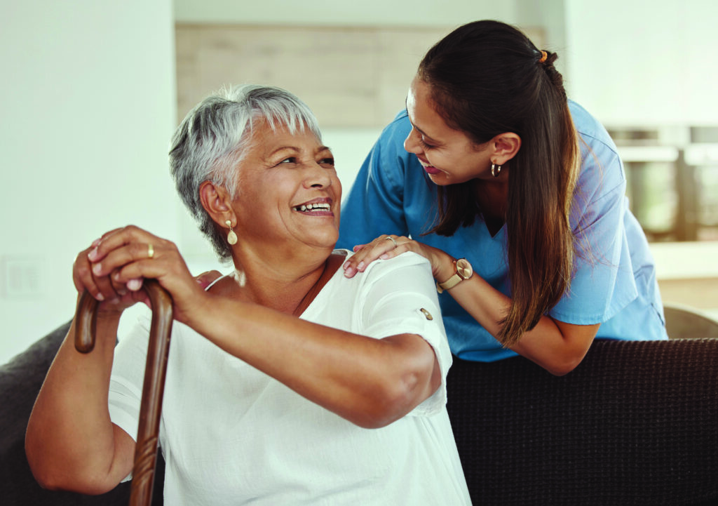 Happy, relax and senior woman with caregiver smile while sitting on a living room sofa in a nursing home. Support, help and professional nurse or healthcare worker helping elderly lady or patient