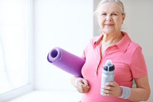 A senior woman stands with her yoga mat and water bottle