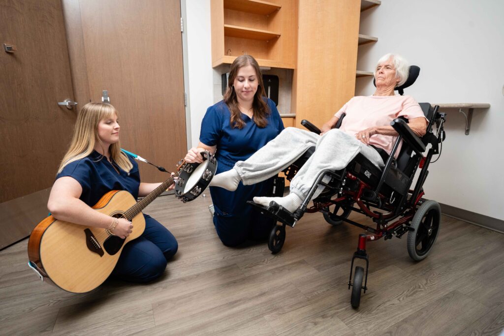 A music therapists sits on the hospital room floor playing a guitar while an speech therapists shakes a tambourine with a patient listening.