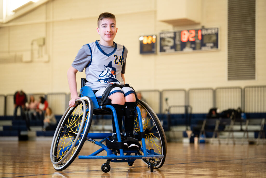 a child in a basketball outfit in a wheelchair poses on a basketball court.