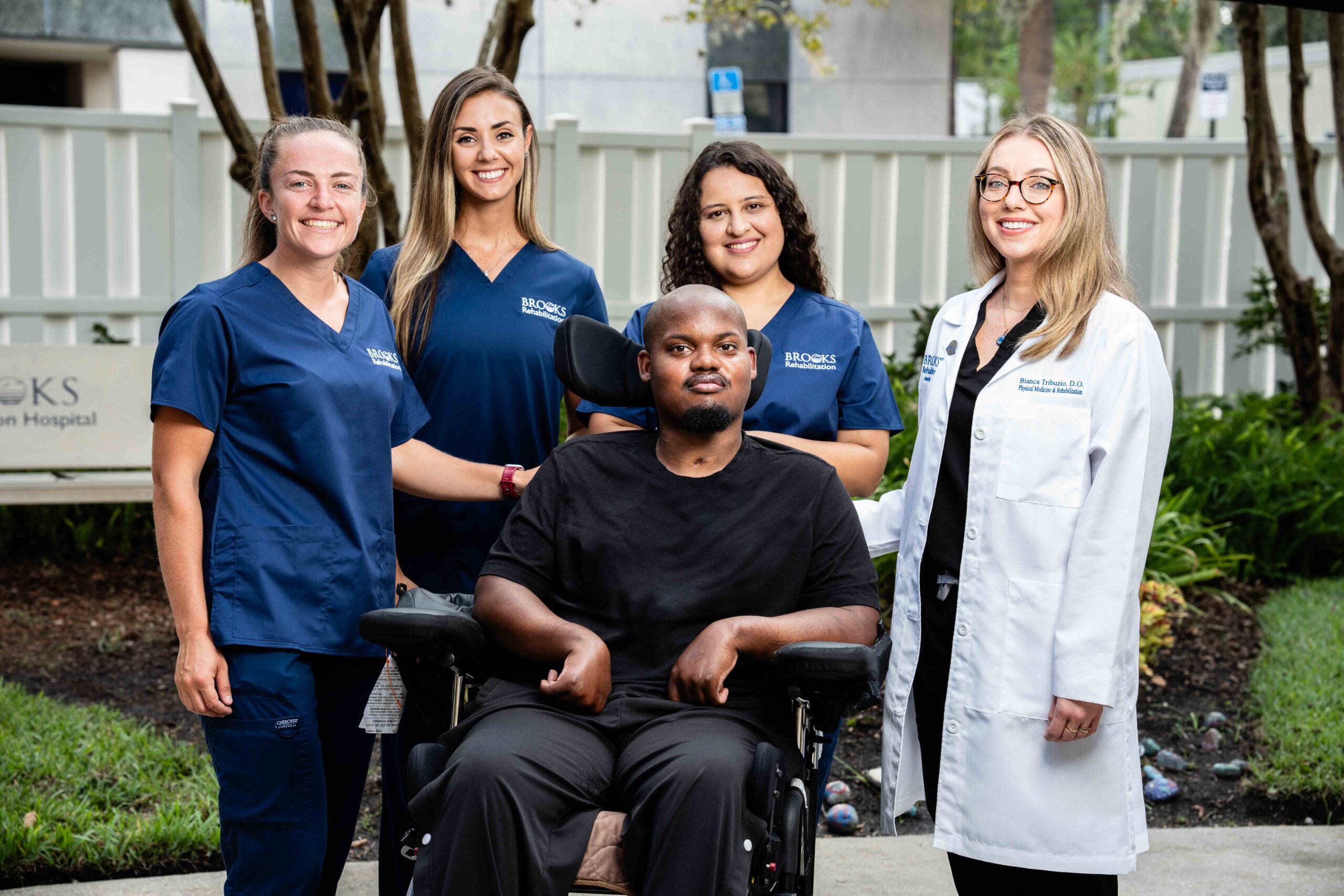 Brooks doctor, nurses and therapists stand around a patient in a a black wheelchair.
