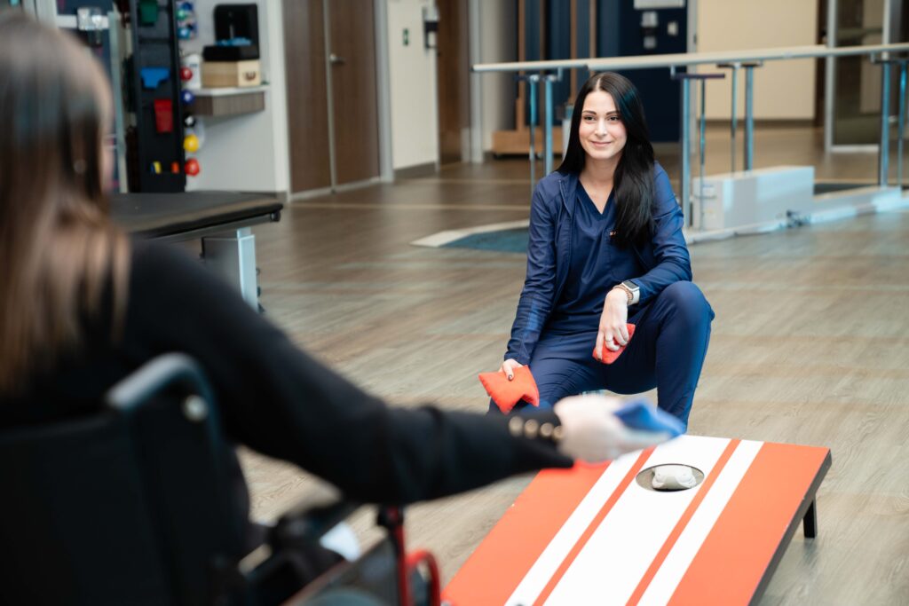 Recreational therapist coaches a patient in a wheelchair on throwing a bean bag into the cornhole box.