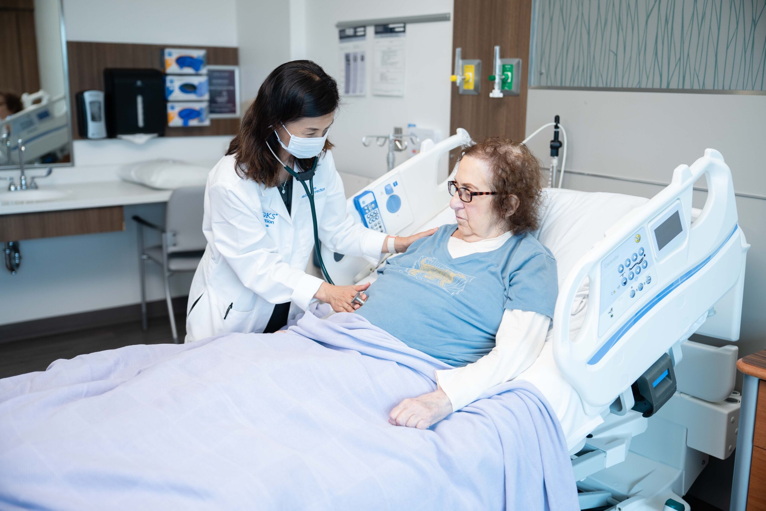 Doctor wearing a mask exams a female transplant patient in their hospital bed.
