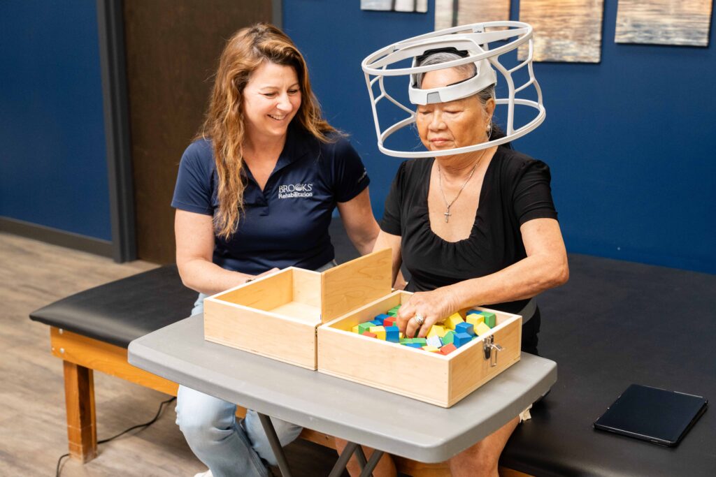 an elderly woman stroke research participant wears a large apparatus on her head while picking up wood blocks of different colors.