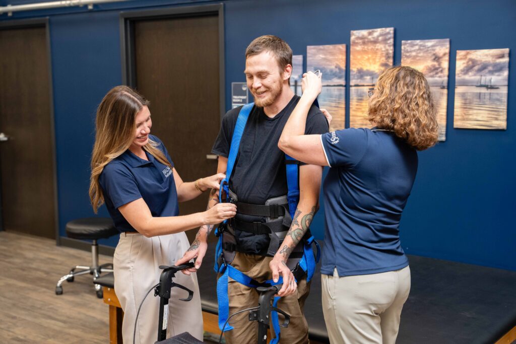 Two brooks research clinicians assist a spinal cord injury study participant into a blue harness for walking tests.