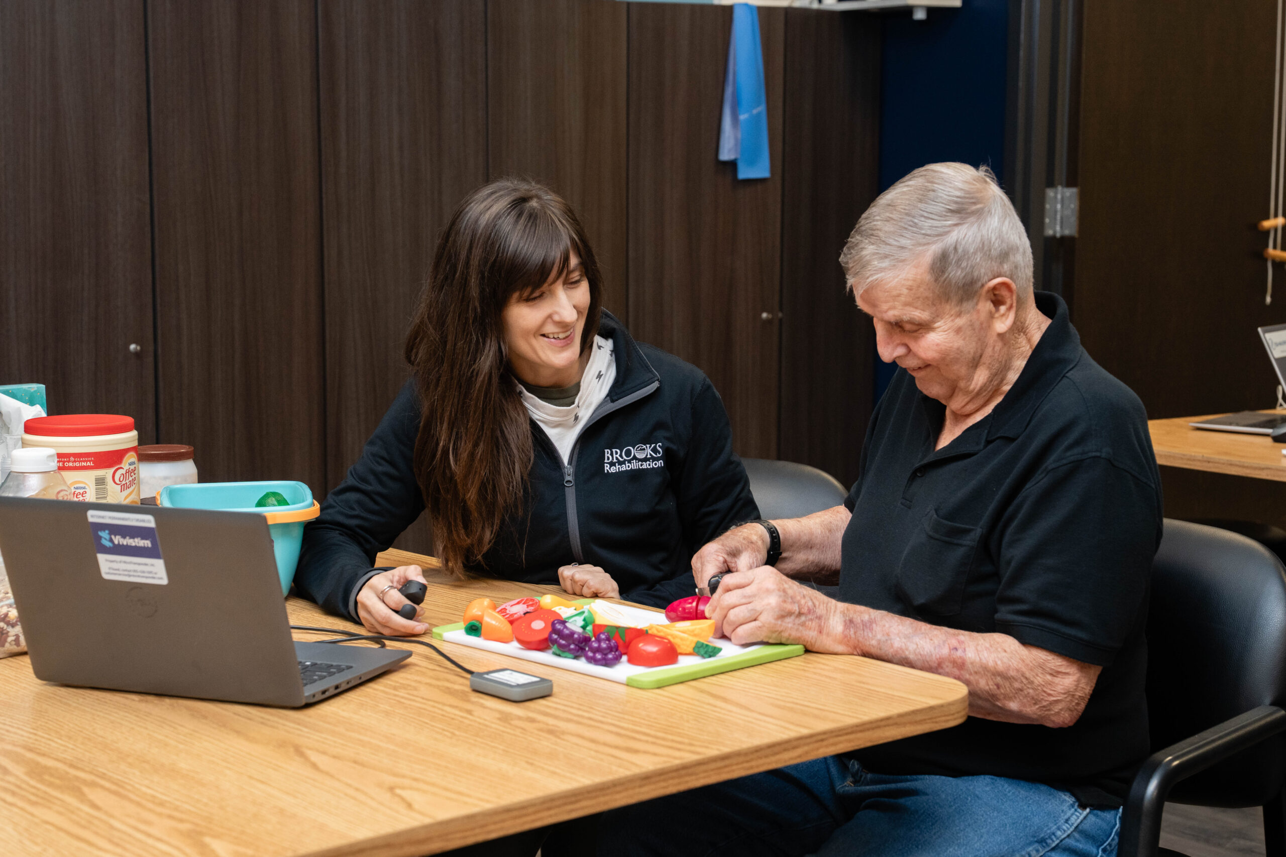 An occupational therapist works with an elderly male patient on cutting vegetables while using the VNS device.