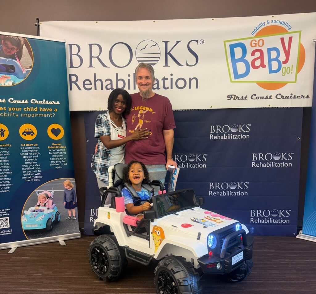 Family smiling in front of GoBabyGo and Brooks Rehabilitation sign along with a child in a toy car