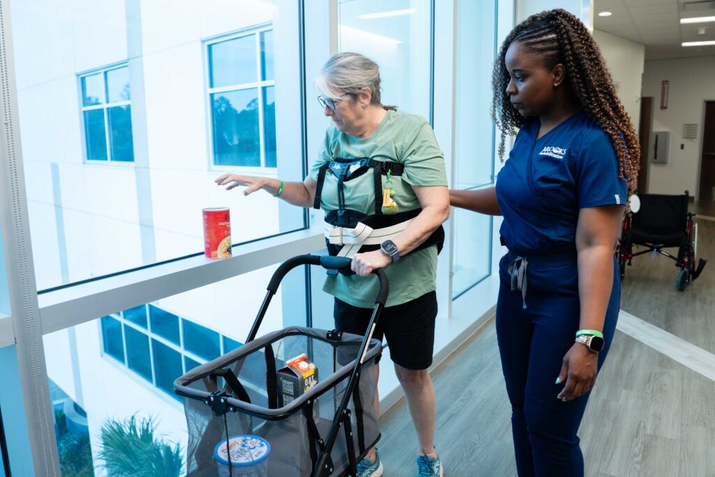 Occupational therapist in blue scrubs walks with an elderly female patient who is pushing a shopping cart and practicing picking up a canned vegetables off a ledge to put in the cart.
