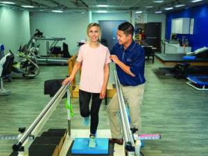 A physical therapists supports a female patient on a balance bar in a clinic gym.