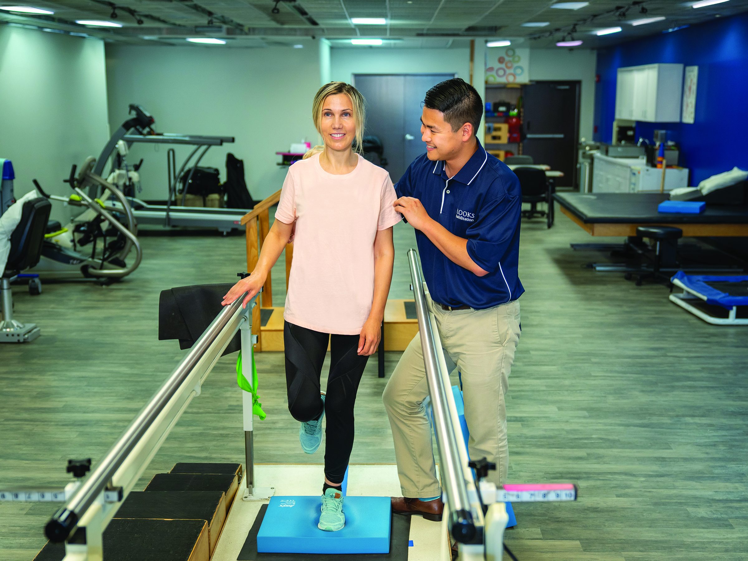 A physical therapists supports a female patient on a balance bar in a clinic gym.